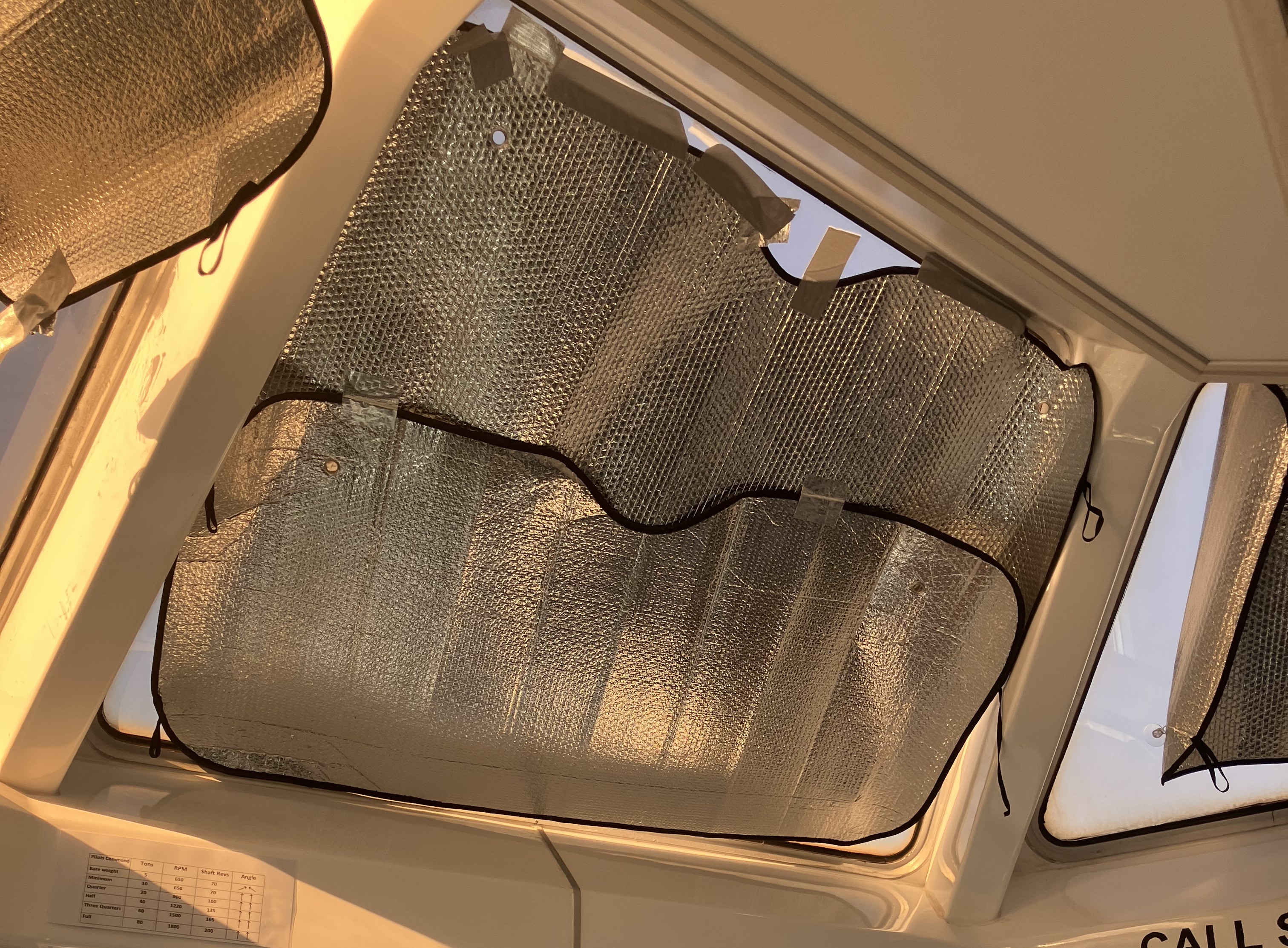 Inside a tugboat wheelhouse, looking up at the angled upper windows. Silver bubble foil car windshield sun shades have been cut to shape, overlapped and held on with strips of grey tape. They cover almost all of the glass, so the only light comes in around the edges and nothing outside can be seen.