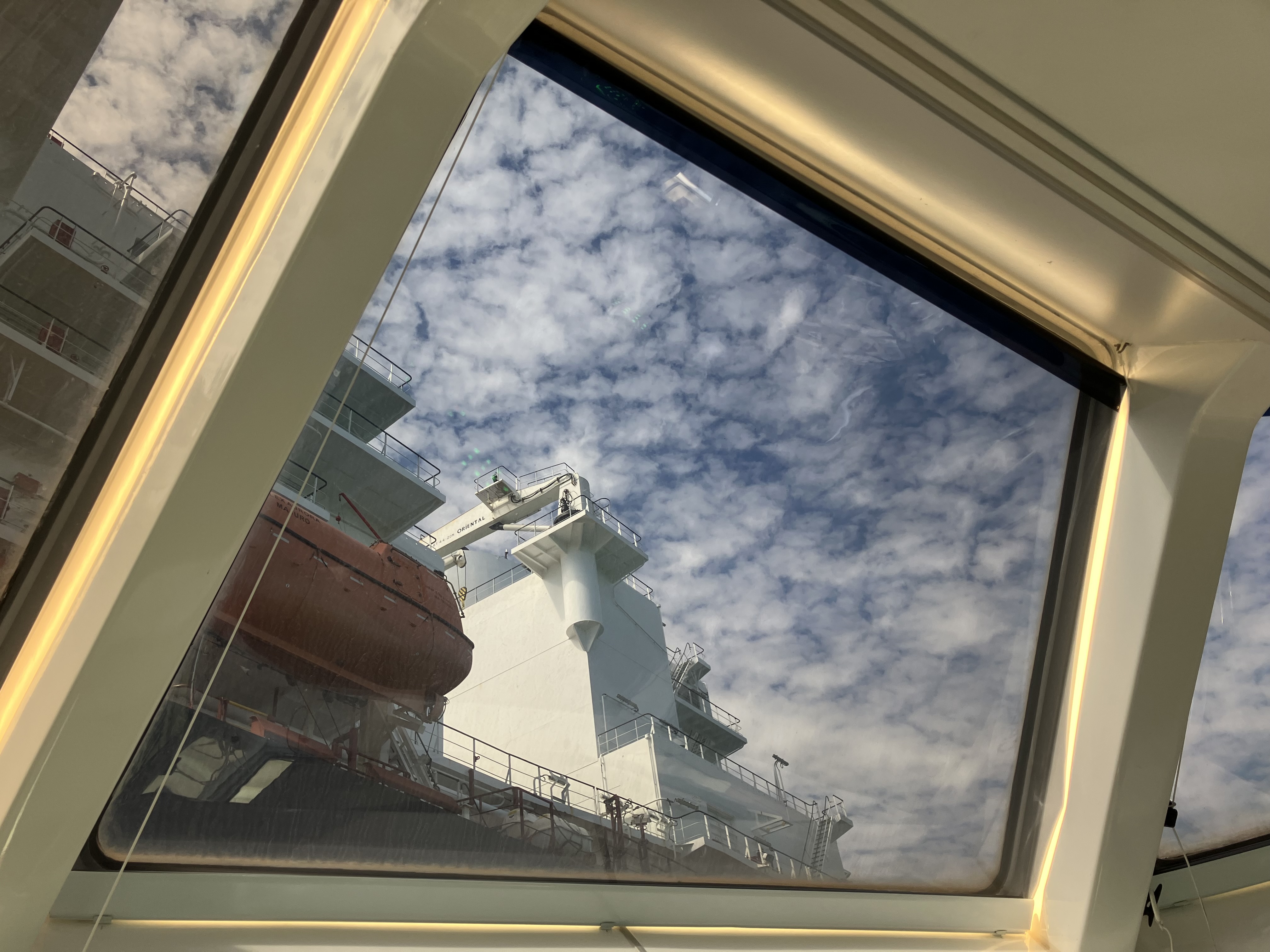 Inside a tugboat wheelhouse, looking up through an angled upper window. The view outside is clear and sharp: a large white ship close alongside, with an orange lifeboat and a deck crane, against a blue sky with scattered cloud.