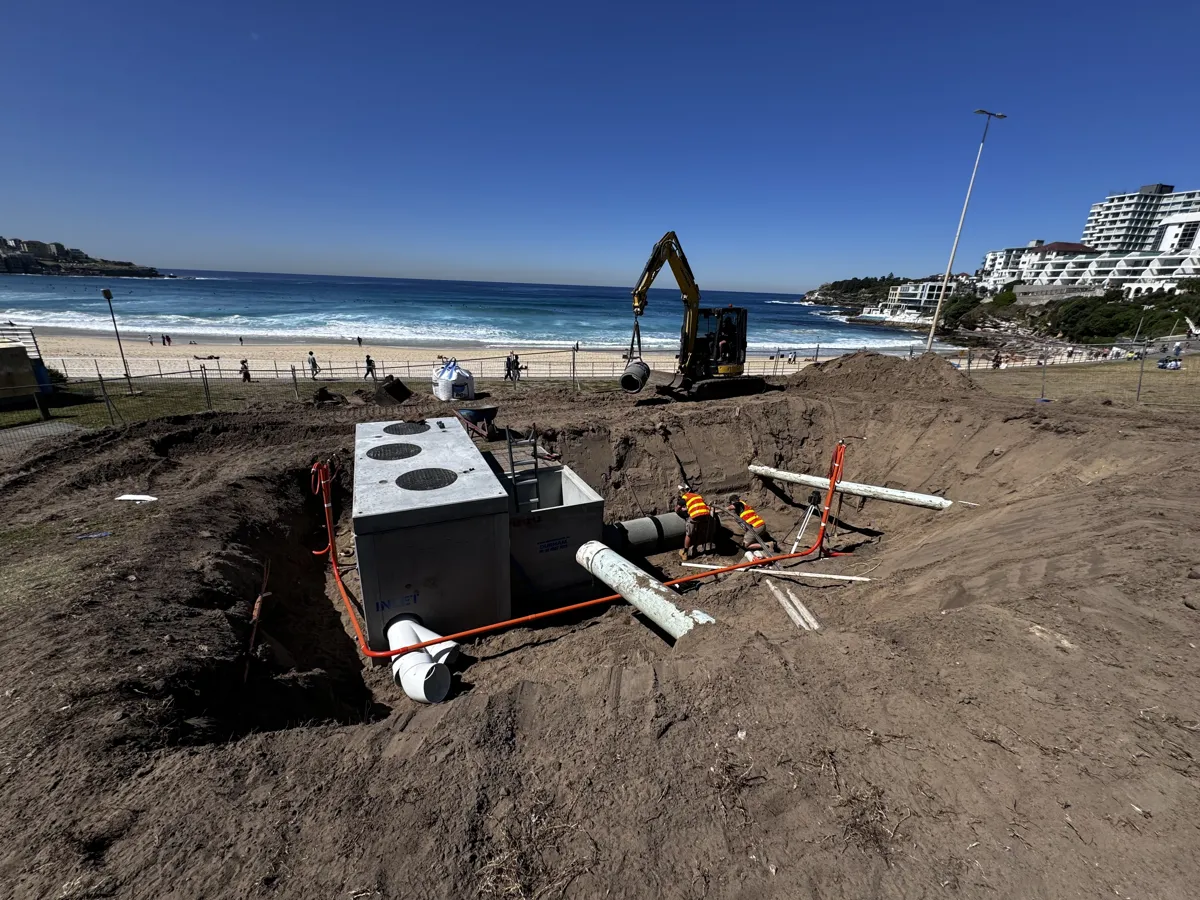 Construction workers in high-visibility vests working on underground pipes and a concrete structure near a beach with buildings in the background under a clear blue sky.