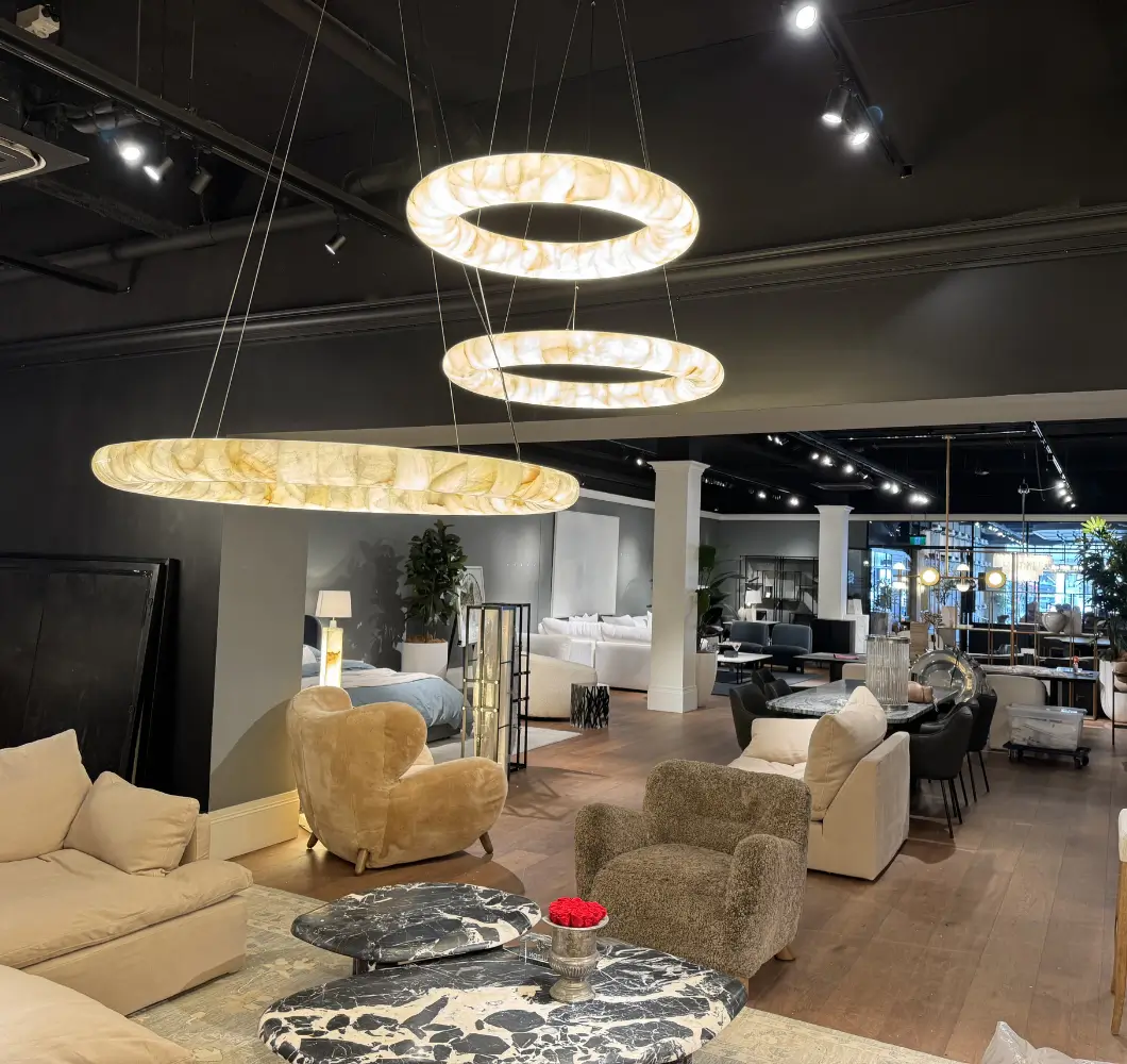Modern furniture showroom with beige and brown armchairs, marble coffee table, and three circular hanging pendant lights.