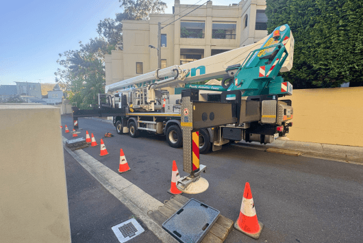 Truck-mounted crane parked on a street with orange traffic cones around it and an open utility cover in front.