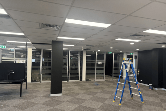 Empty office space with carpeted floor, white fluorescent ceiling lights, glass partitions, and a blue step ladder near a dark wall.