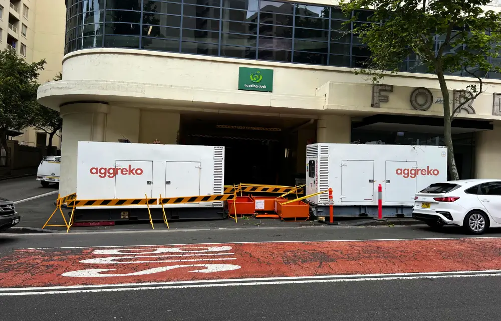 Two large white Aggreko generators placed on a street in front of a building's loading dock, with orange and black barricades surrounding them.