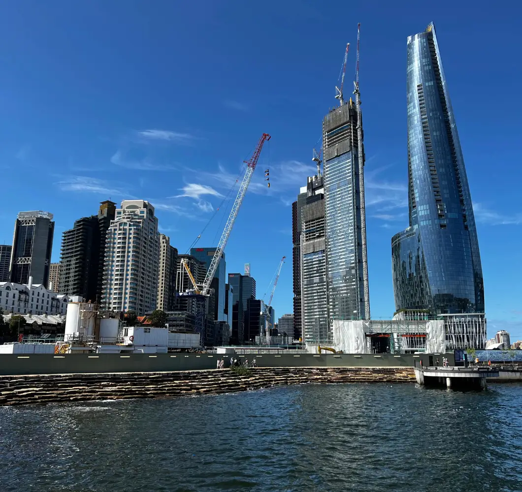 City skyline featuring modern high-rise buildings under construction with cranes beside a waterfront under a clear blue sky.