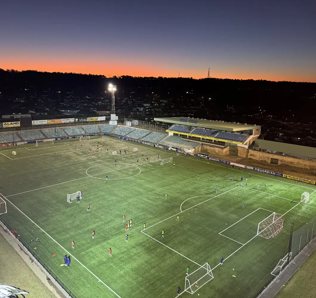 Aerial view of a soccer stadium with players practicing on the field at dusk under floodlights.