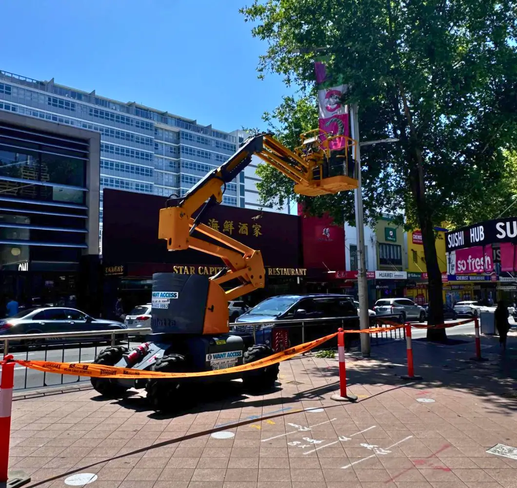 Yellow articulated boom lift elevated on a sidewalk near a restaurant, with safety tape and orange cones around the area.