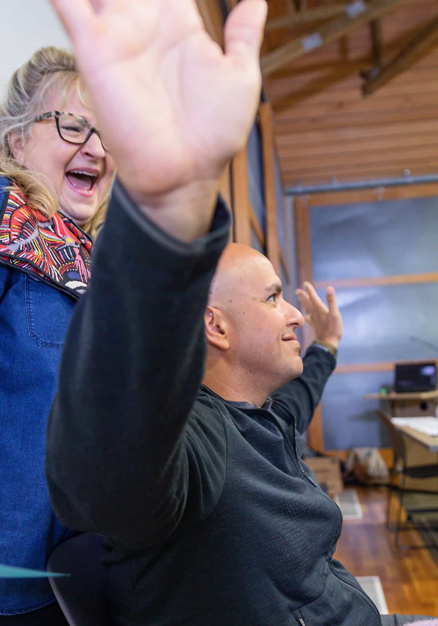 man raising his arms up and laughing; woman in background smiling
