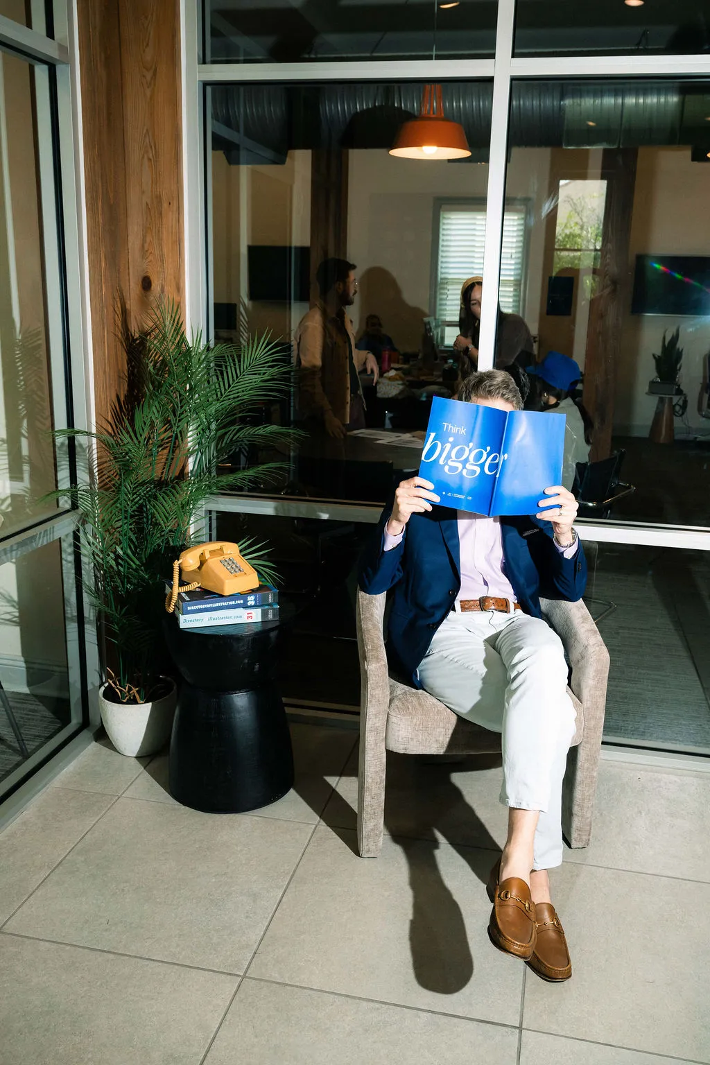 Taylor seated in a lobby chair holding a blue book in front of his face