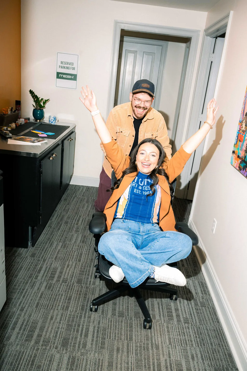 Two team members with their arms raised in an office doorway on a rolling chair