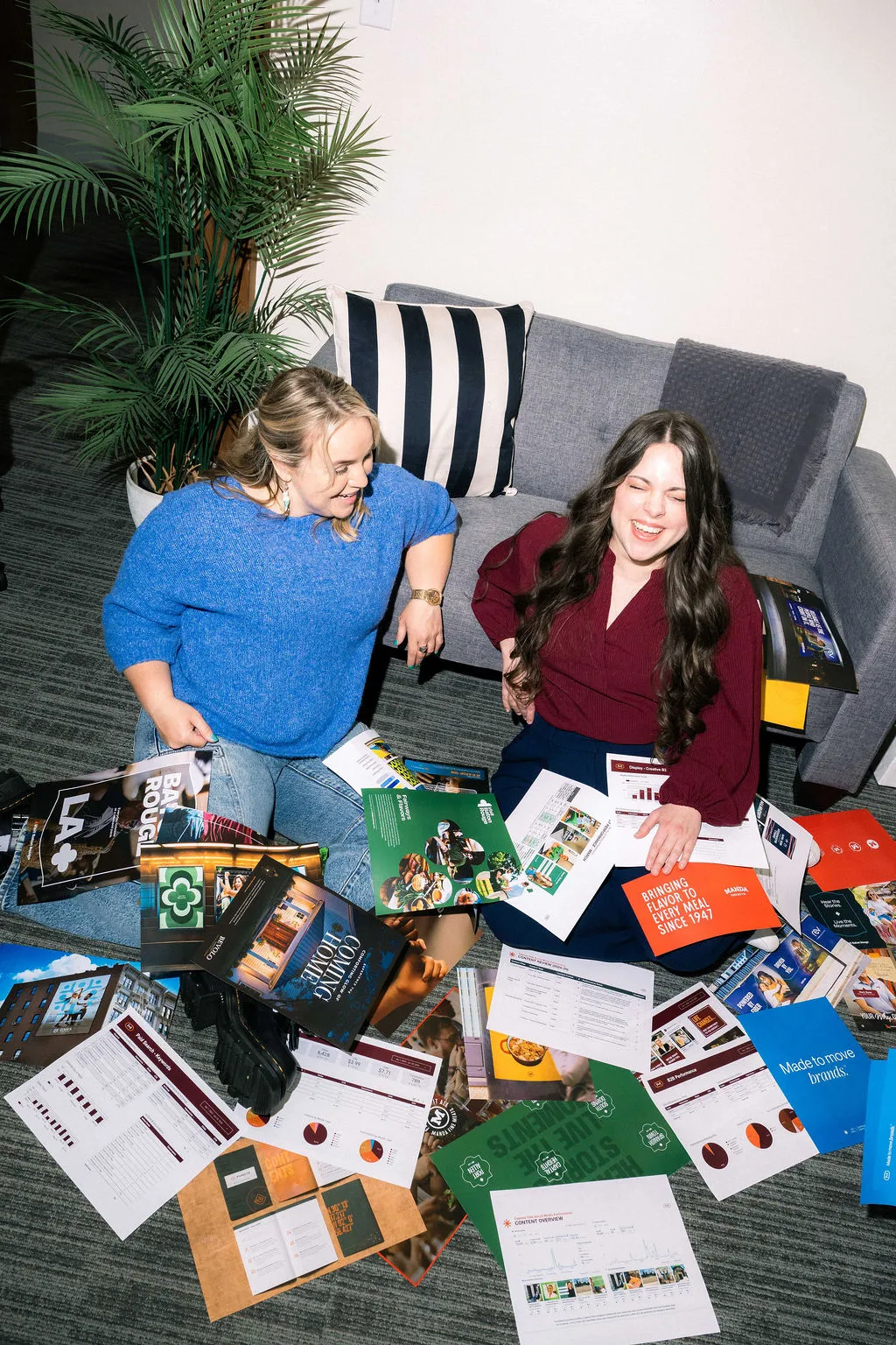 Two women sitting on the floor surrounded by printed brochures and packaging samples
