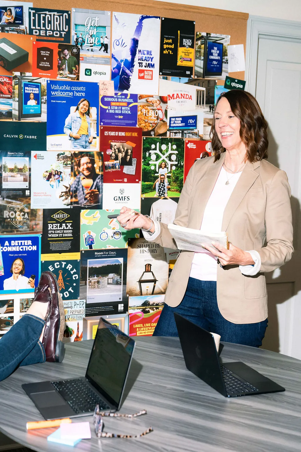 A woman presenting beside a wall covered in printed client work, with a colleague's feet propped on the table