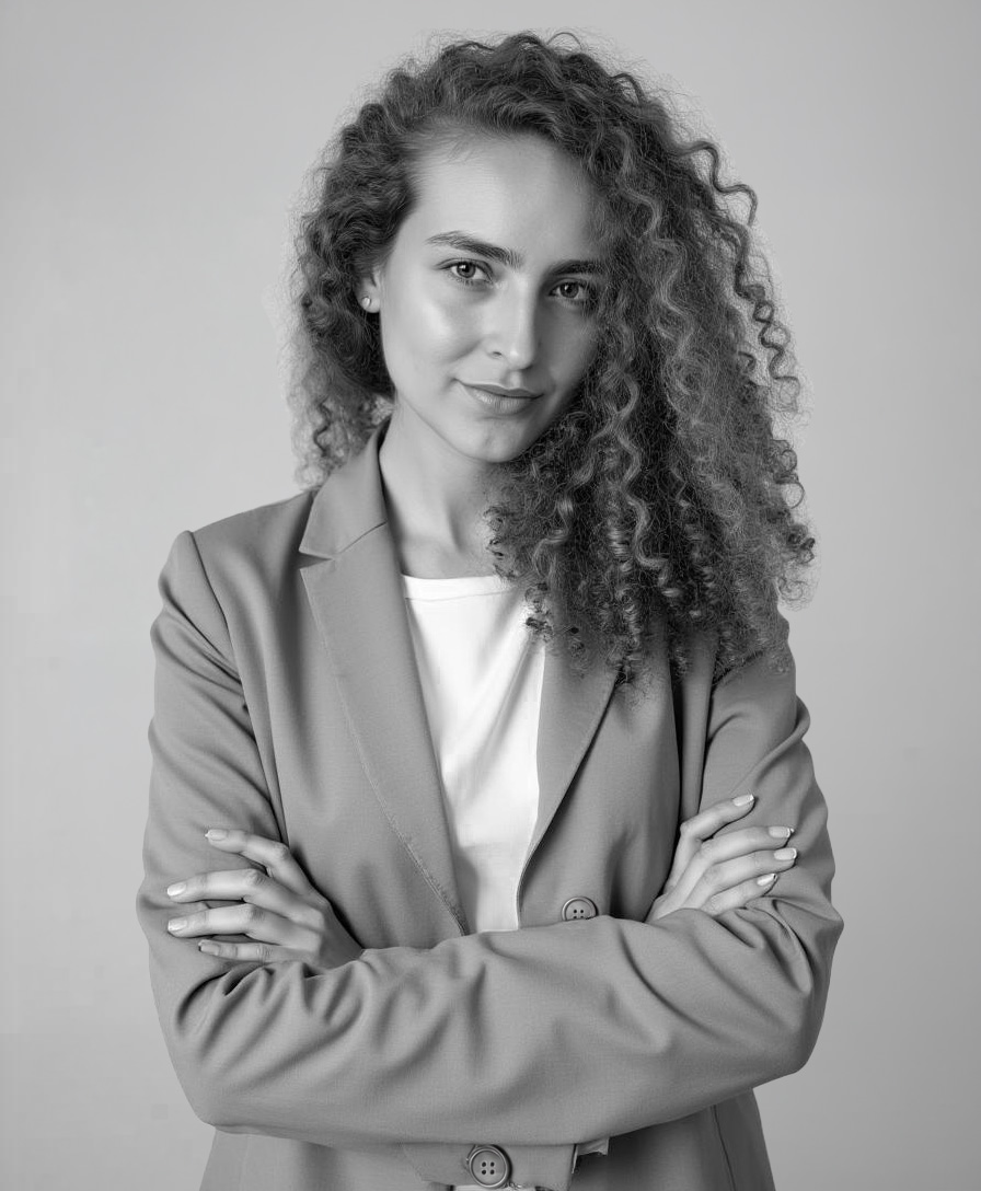 Confident woman with long curly hair wearing a blazer and white top, standing with arms crossed.