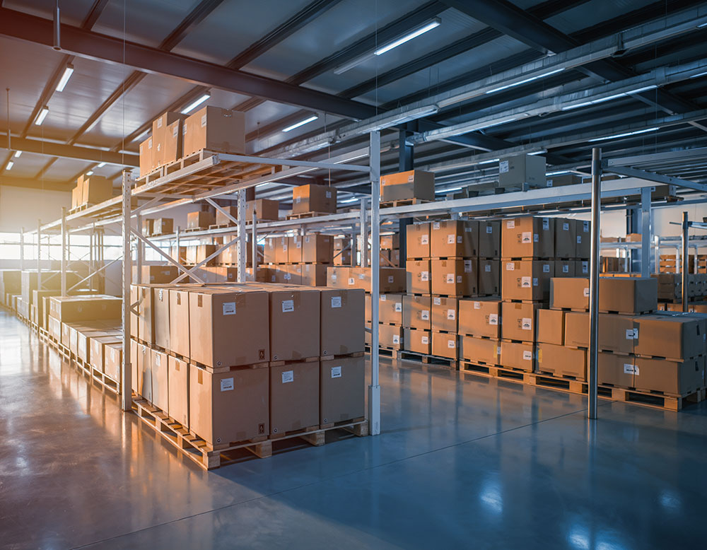 Warehouse interior with rows of stacked cardboard boxes on metal shelving and pallets, illuminated by overhead lights and sunset.