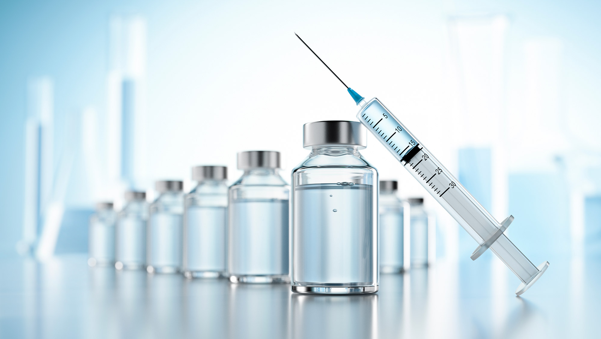Row of vaccine vials with a syringe resting diagonally on the front vial in a bright laboratory setting.