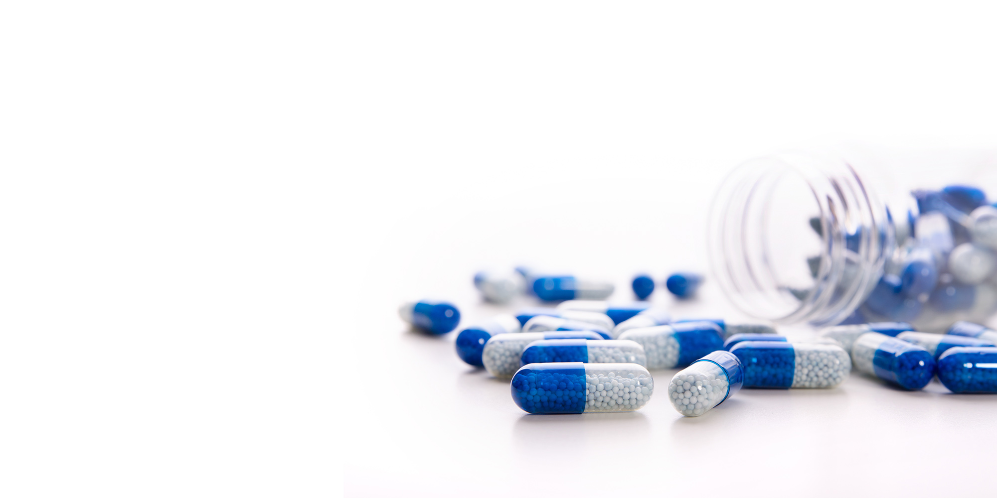 Blue and white capsules scattered on a white surface with a clear, tipped-over pill bottle in the background.