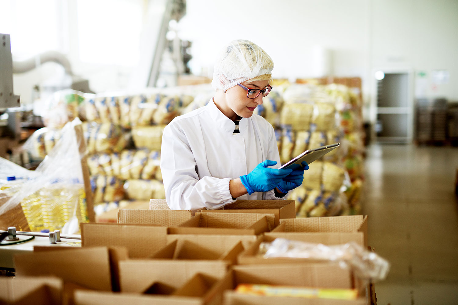 Woman in protective clothing and gloves checking a tablet in a food packaging facility with stacked packaged goods in the background.