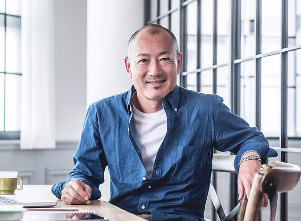 Smiling man in blue shirt sitting comfortably at a wooden table in a bright office with large windows.