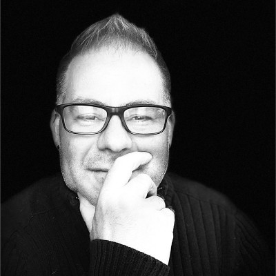Black and white portrait of a man wearing glasses with short hair and a goatee, resting his chin on his hand.