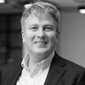 Smiling man with light hair wearing a suit jacket and white shirt in an indoor setting.