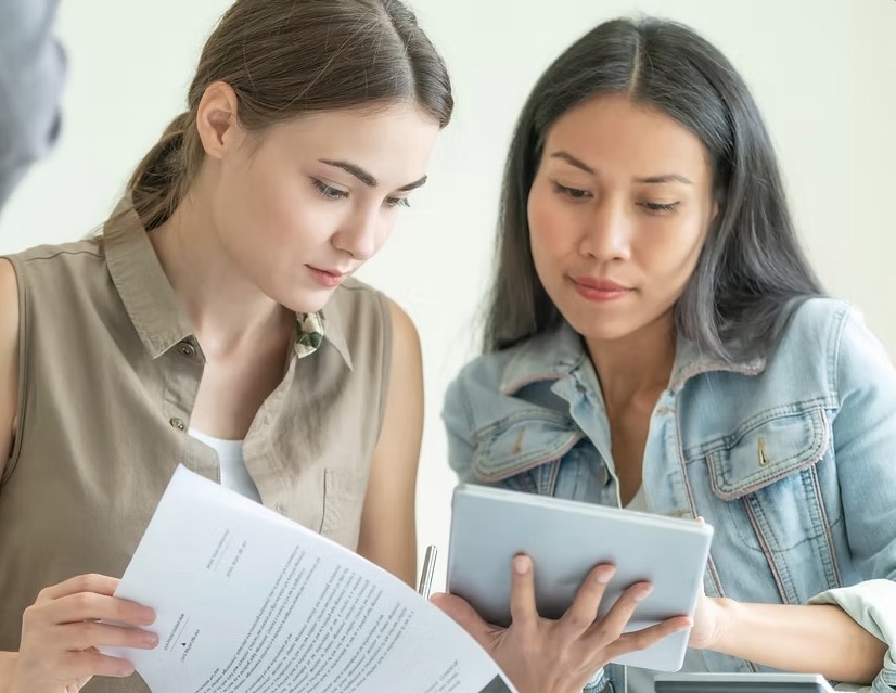 Two women focused on reviewing documents, one holding papers and the other using a tablet.
