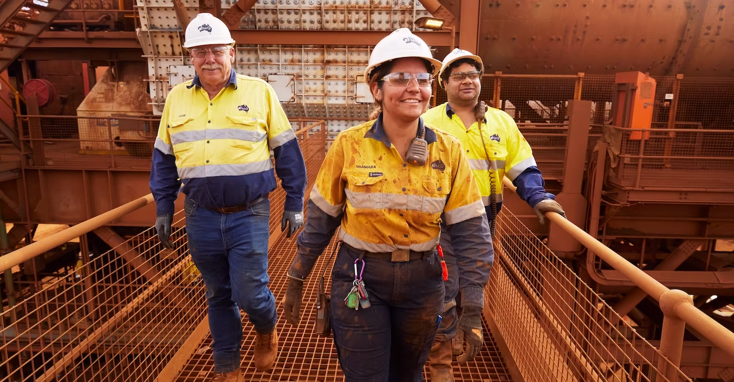 Three industrial workers wearing hard hats and high-visibility shirts walking on a metal walkway in a factory setting.