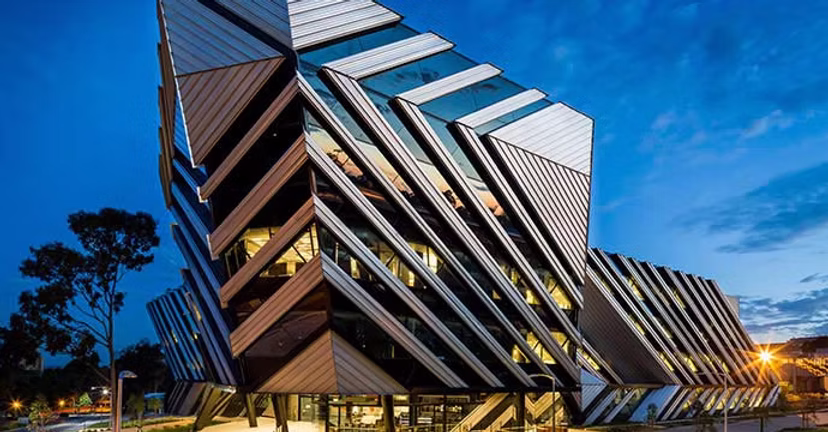 Modern building with geometric, angular metal panels illuminated at dusk with a clear blue sky in the background.