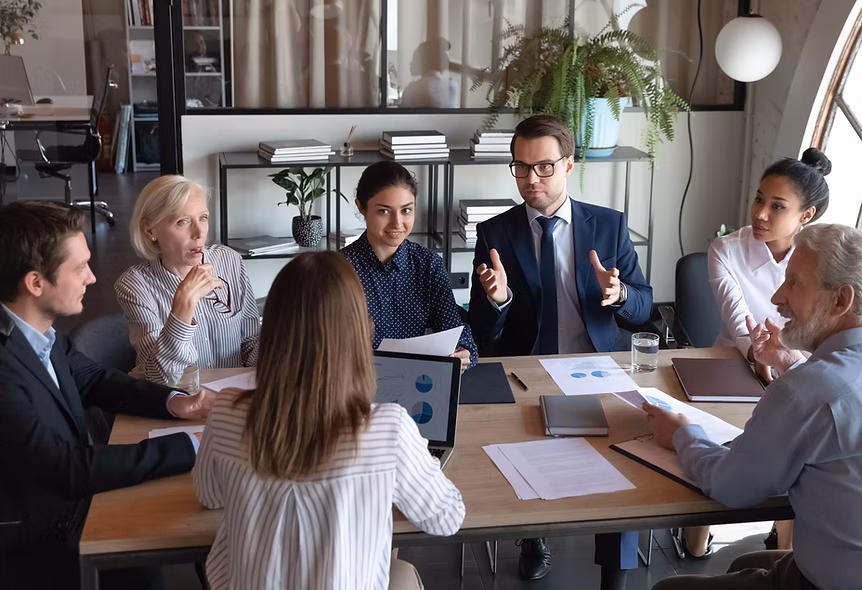 Six diverse professionals engaged in a discussion around a wooden table with documents and laptop.