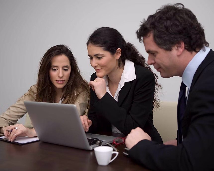 Three business professionals, two women and one man, collaborating and viewed focused on a laptop at a conference table with a coffee cup.
