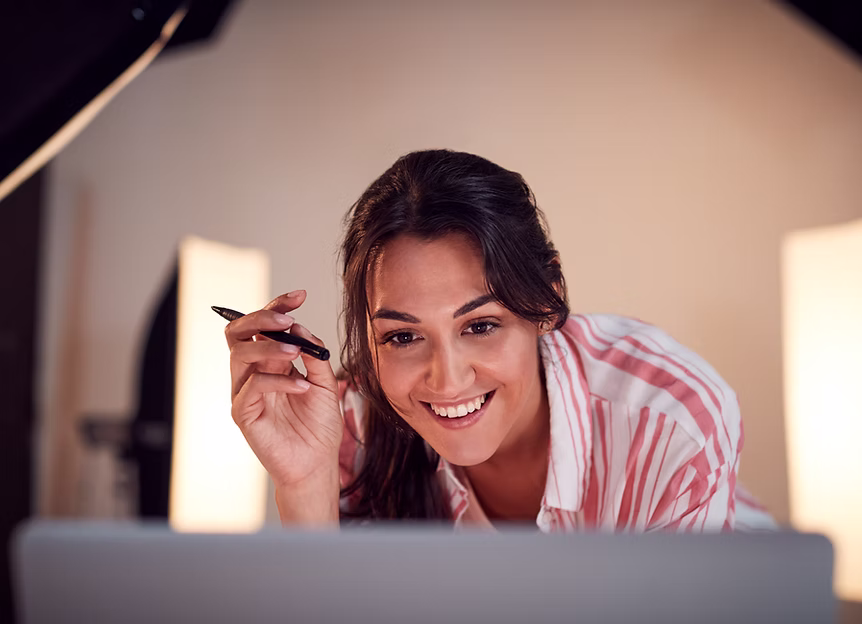 Smiling woman holding a pen and looking at a laptop screen in a softly lit room.