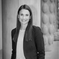 Smiling woman with long straight hair wearing a blazer and sweater, standing indoors near a wall with circular patterns.