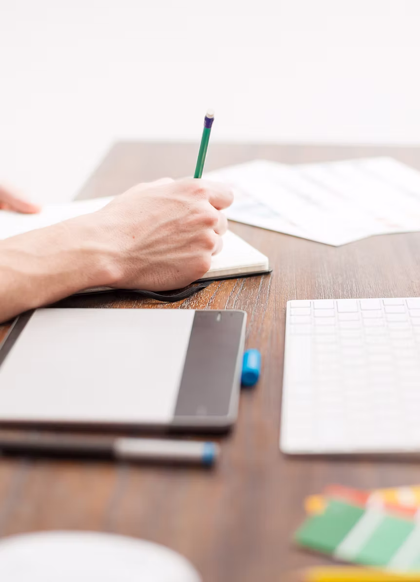 Person writing with a green pencil on an open notebook on a wooden desk with a keyboard, graphics tablet, and color swatches nearby.