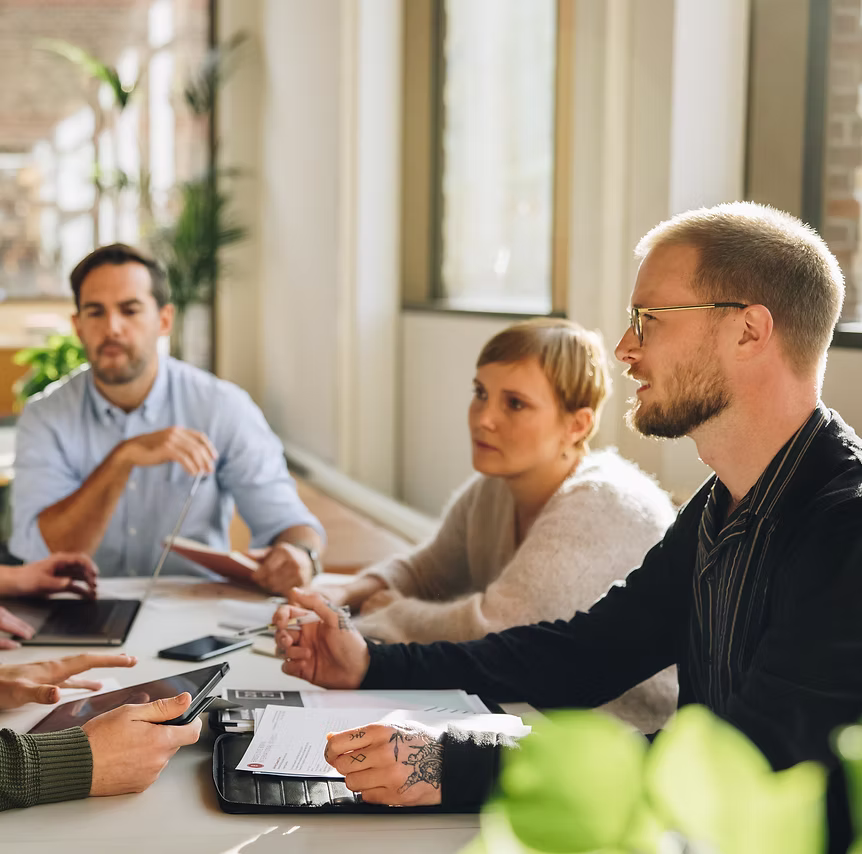 Group of four adults engaged in a discussion around a table with documents and electronic devices in a bright office.