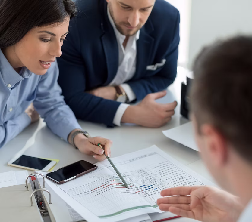 Three people in a business meeting discussing a printed chart with graphs on a table.