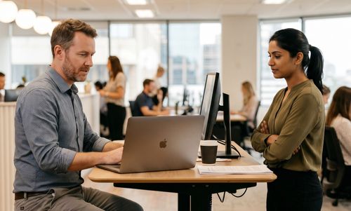 Two coworkers collaborating at a standing desk in a modern office with computers and paperwork.