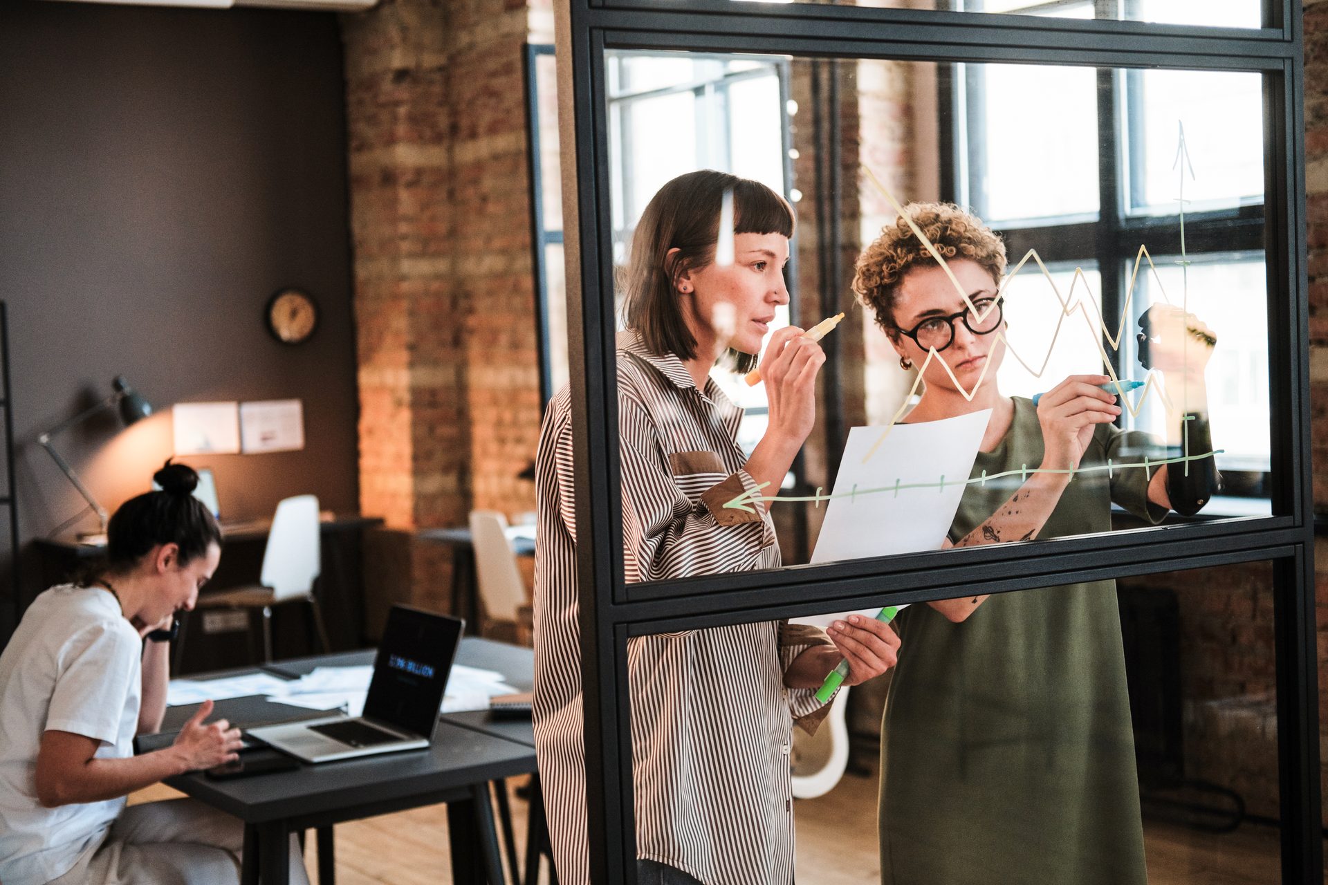 Two women collaborating on a line graph drawn on a glass wall while another person works on a laptop in the background in a modern office.