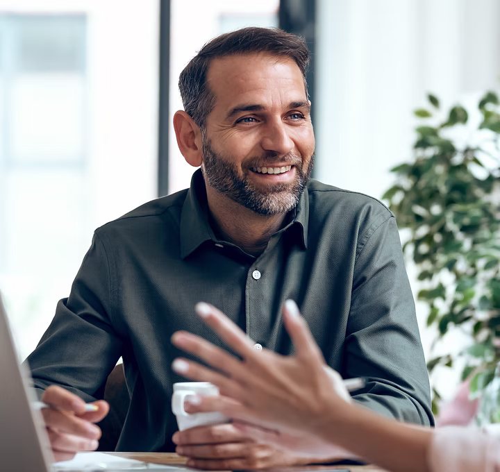 Smiling man with beard in a dark green shirt holding a coffee cup during a conversation in a bright office.