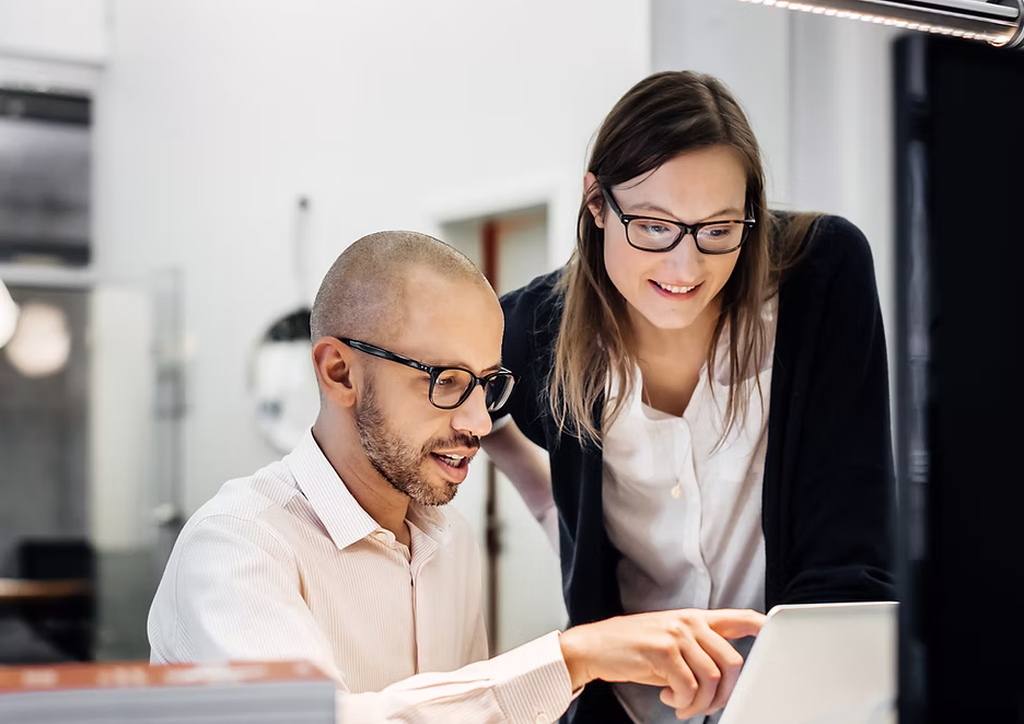 Two colleagues wearing glasses smiling and working together on a laptop in a modern office.