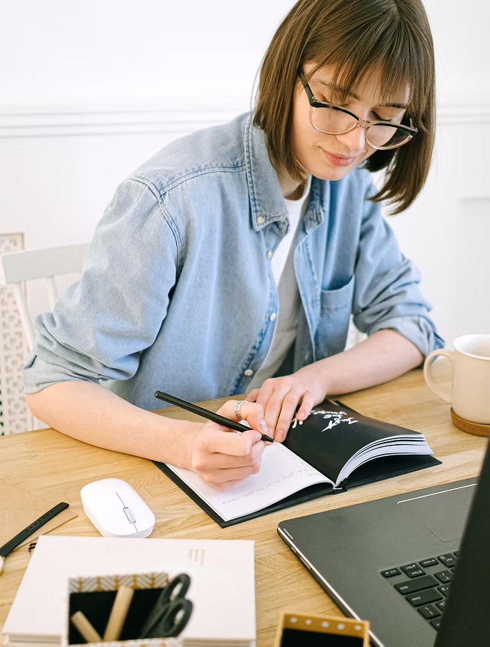 Young woman with glasses writing in a notebook at a wooden desk with a laptop, mouse, and mug nearby.