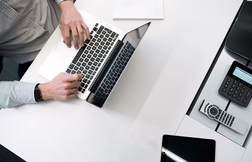 Person typing on a laptop on a white desk next to a remote control, calculator, and a tablet.