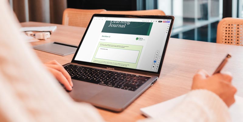 Person writing notes on paper with a laptop open to a digital learning journal on a wooden table.
