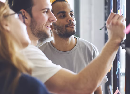 Three people collaborating and placing sticky notes on a glass wall during a brainstorming session.