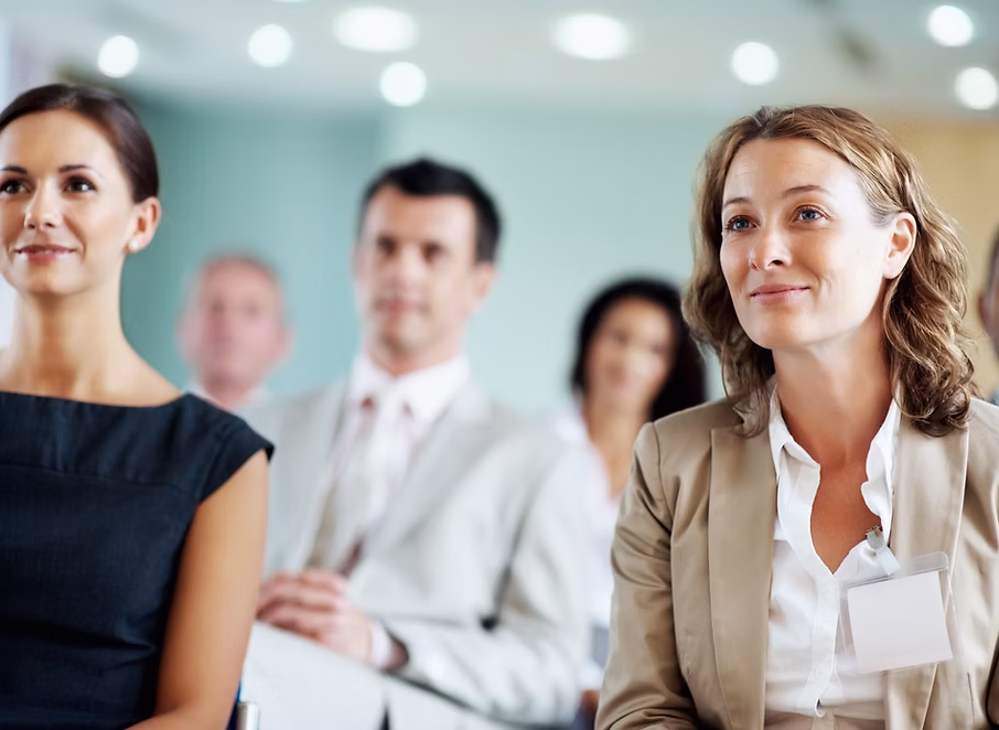 Business professionals attentively listening at a conference with blurred audience in the background.