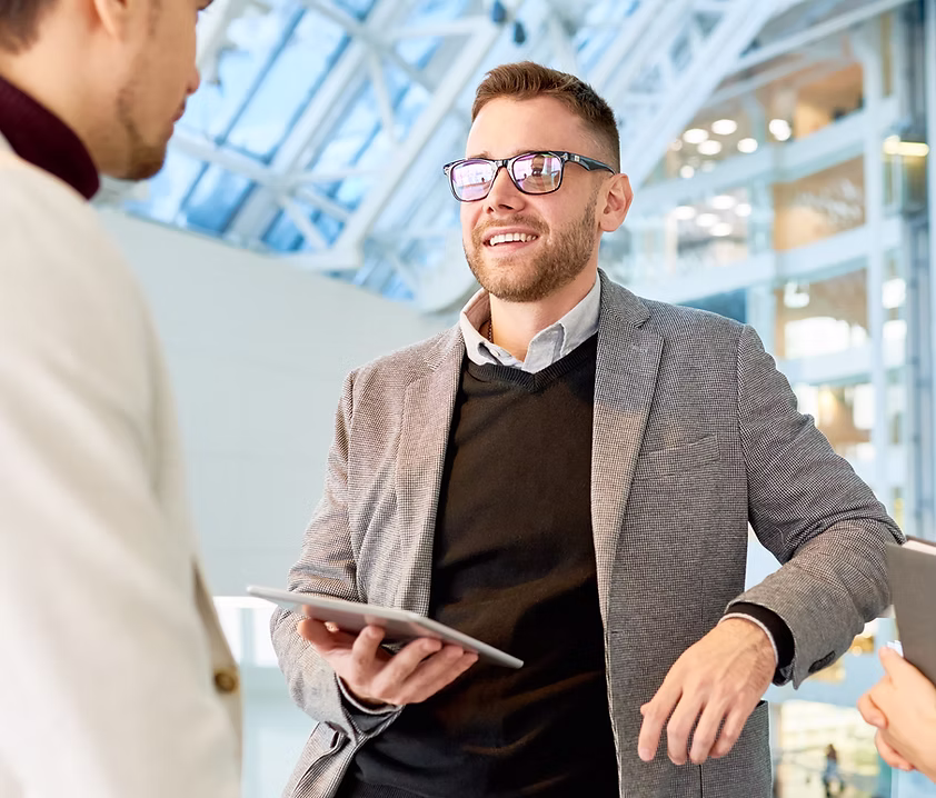 Smiling man wearing glasses and a gray blazer holds a tablet while talking to two people in a modern office space.