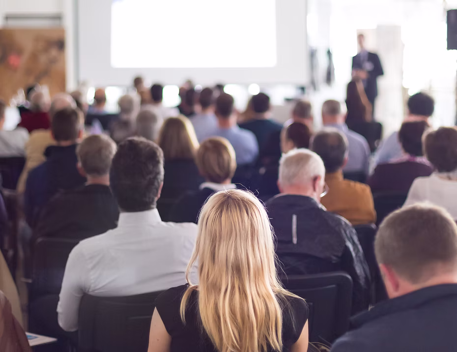 Audience seated facing a presenter giving a business presentation in a conference room.