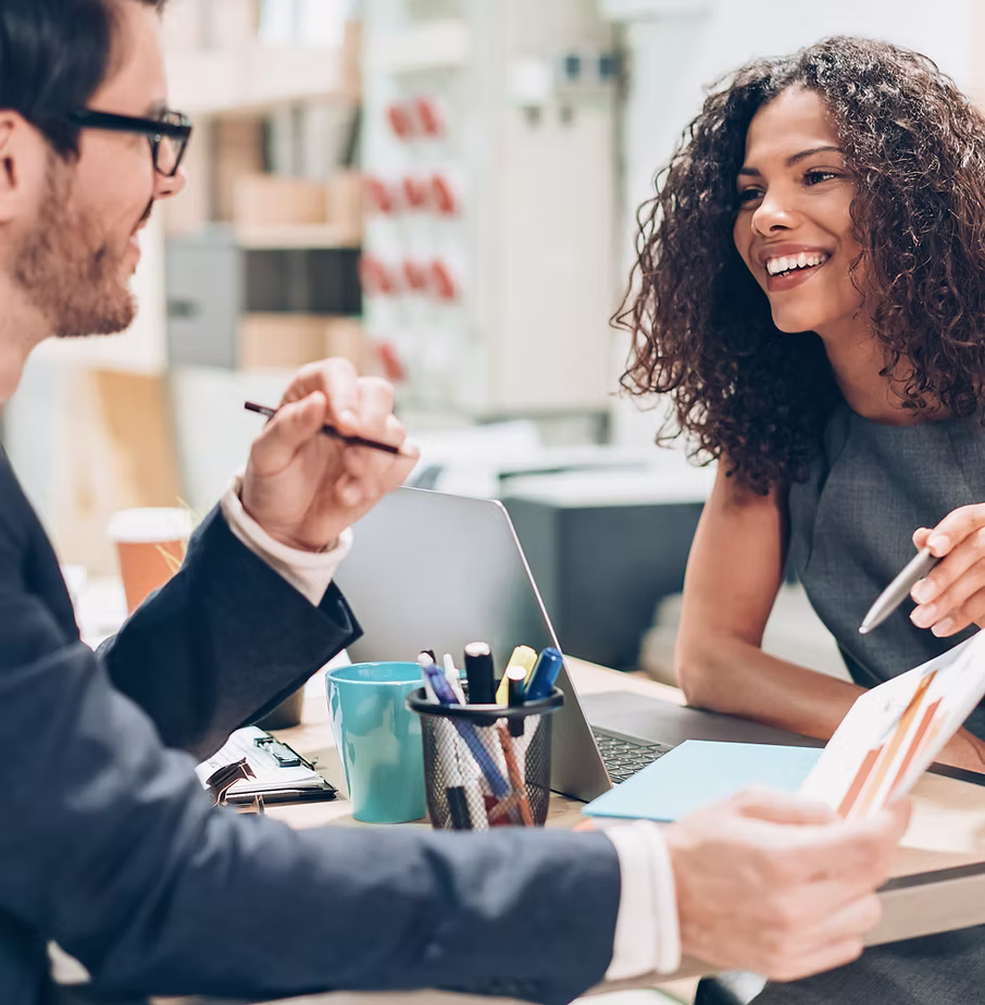 Two young business colleagues engaged in a discussion at a desk with a laptop, papers, and stationery.
