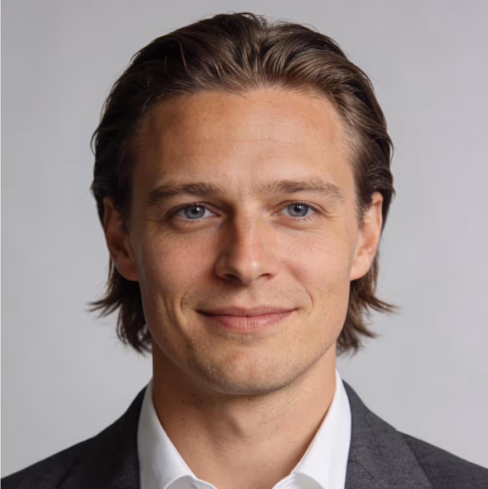 Smiling young man with light blue eyes and slicked-back brown hair wearing a white shirt and dark blazer.