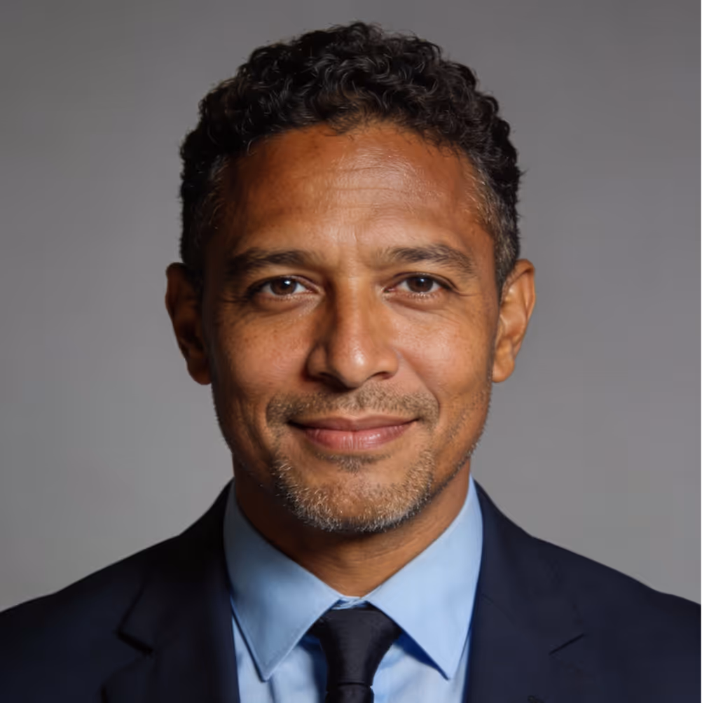 Smiling man with curly hair wearing a dark suit, light blue shirt, and dark tie against a gray background.