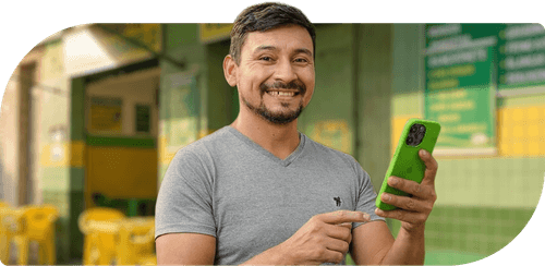 Smiling man in gray shirt holding and pointing at a green smartphone outdoors near yellow chairs.