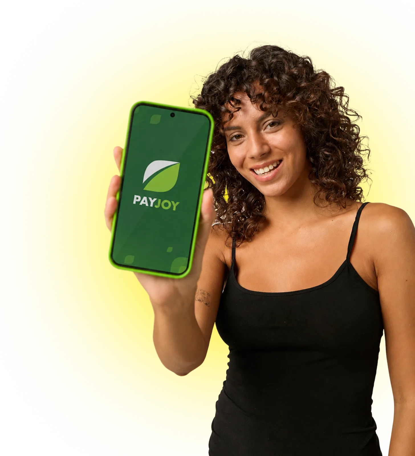 Smiling woman with curly hair holding phone showing PayJoy logo on a green screen, yellow background.