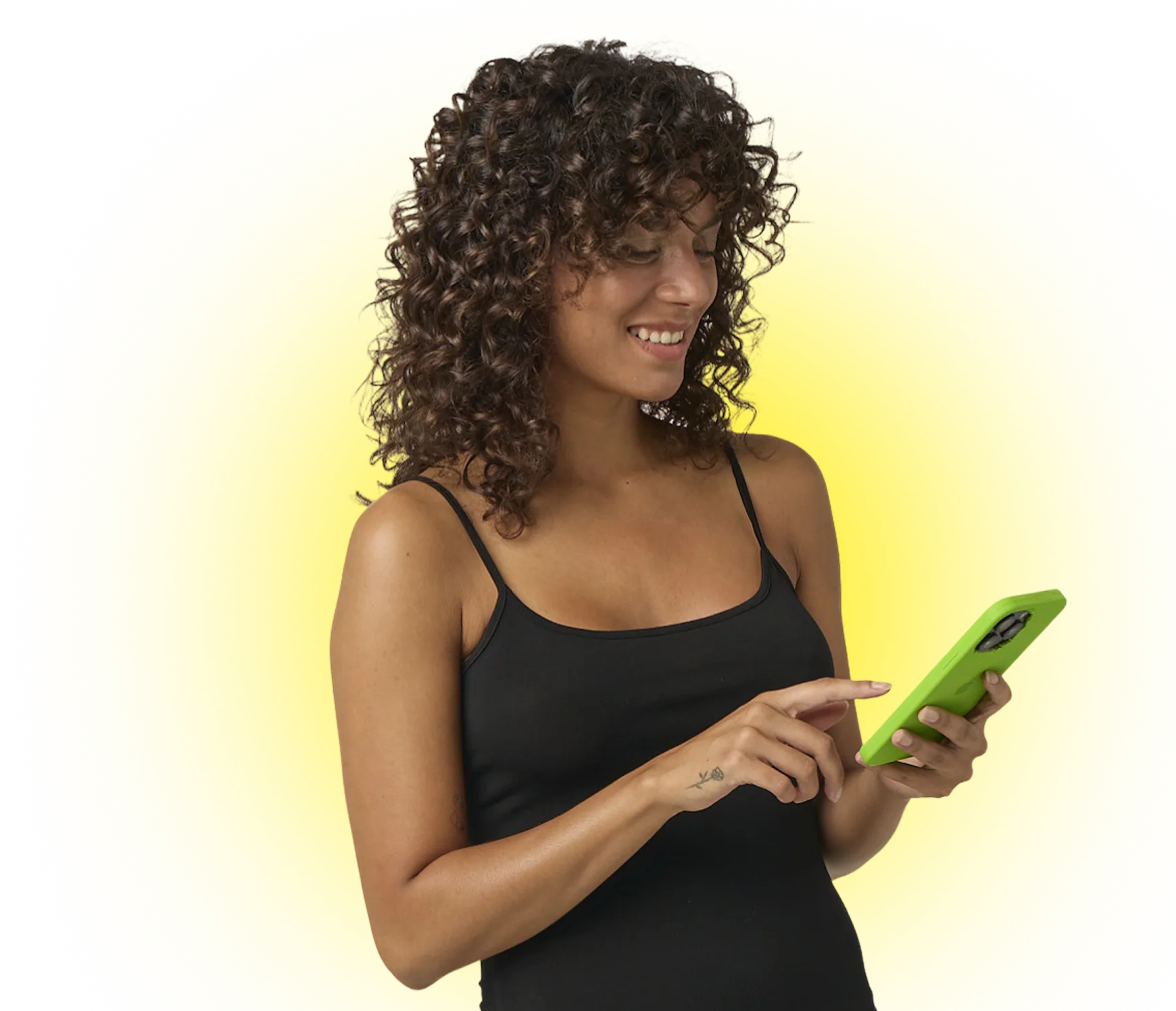 Smiling woman with curly hair in black tank top using a green smartphone against a yellow background.
